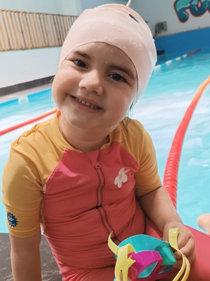 Mayosuyla havuz kenarında oturan bir çocuk elinde deniz gözlüğü tutuyor