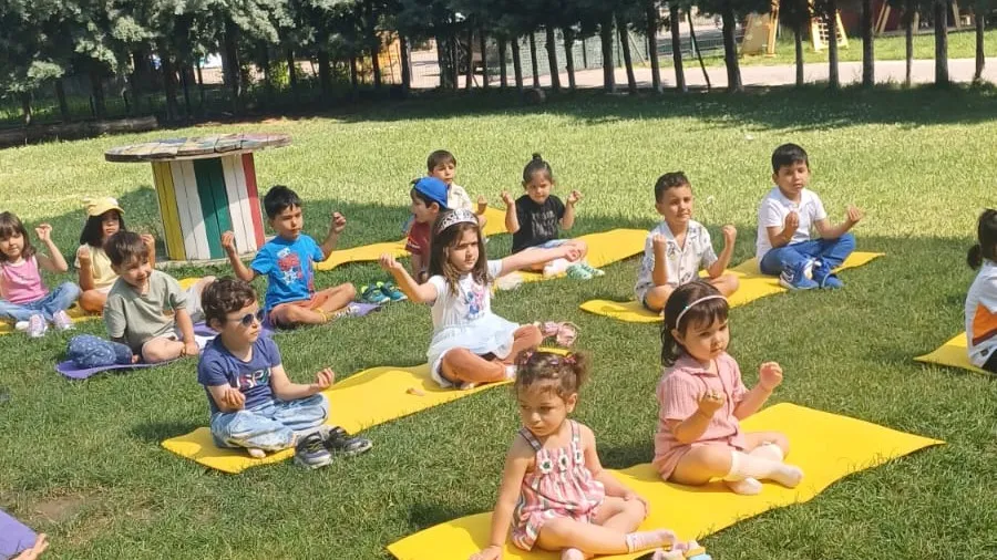 Bahçede sarı minderlerde oturan çocuklar, sabah çemberinde öğretmenin yönergesini el hareketleriyle tekrarlıyor