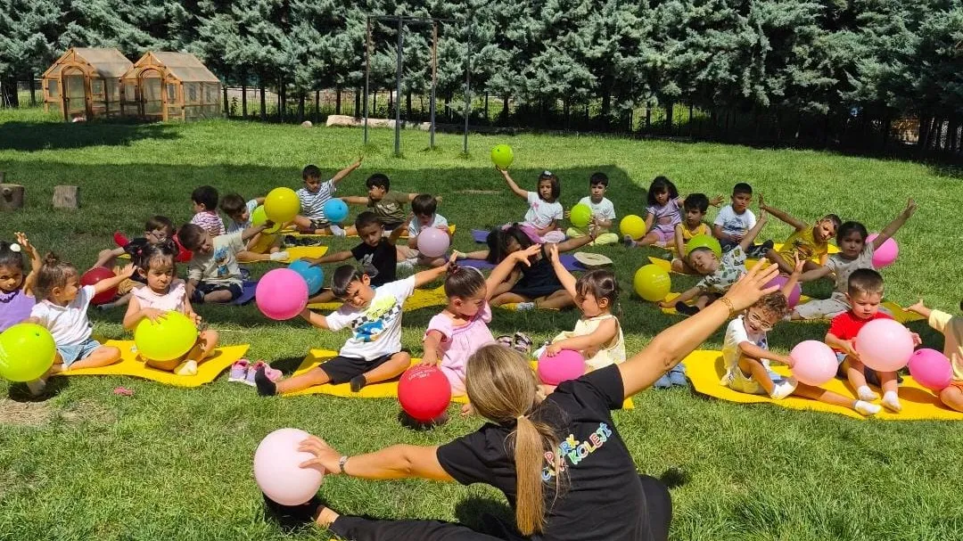 Bahçede minderler üzerinde balonlarla hareket çalışması yapan çocuklar ve öğretmenleri