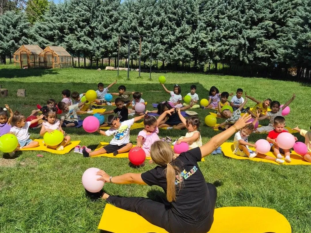 Bahçede minderler üzerinde toplarla koordinasyon çalışması yapan çocuklar ve öğretmenleri